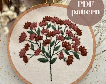 May include: A floral embroidery pattern in a wooden hoop. The design features a stylized flower with brown and pink petals, green leaves, and a brown stem. The text "PDF pattern" is in a circle in the upper right corner.