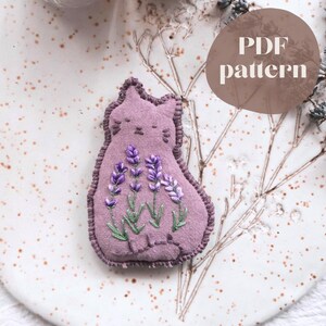 Peut inclure: Un objet en tissu en forme de chat de couleur lavande avec des brins de lavande brodés. Le chat a un contour cousu et les yeux fermés. Un cercle marron avec le texte "PDF pattern" est en haut à droite. L'objet est sur une surface blanc cassé mouchetée.