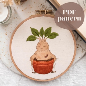 May include: Embroidered hoop art featuring a whimsical mandrake plant with green leaves and a smiling face. The mandrake is in a terracotta-colored pot. The text "PDF pattern" is in a brown circle.