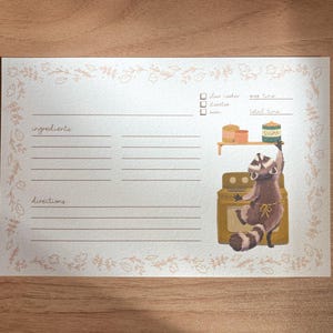 May include: A printable recipe card with a raccoon illustration. The card has a floral border and sections for ingredients and directions. The raccoon is wearing a chef's hat and holding a jar of jam. The text on the card reads "flour, sugar, oven, prep time, total time".