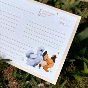 Peut inclure: Une fiche de recette avec un fond bleu clair et une bordure beige. La carte pr&eacute;sente une illustration &agrave; l'aquarelle de deux poulets et une abeille. Le texte sur la carte comprend des options pour la mijoteuse, la cuisini&egrave;re et le four.