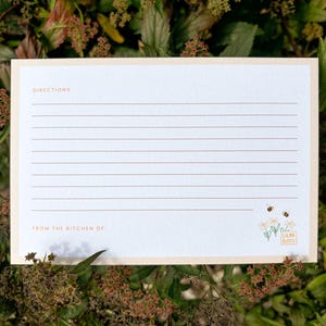Peut inclure: Une carte de recette avec un fond cr&egrave;me et une bordure p&ecirc;che clair. La carte comporte des lignes horizontales pour l'&eacute;criture, avec les mots "DIRECTIONS" et "FROM THE KITCHEN OF" imprim&eacute;s en marron clair. Une petite illustration florale et d'abeilles est dans le coin inf&eacute;rieur droit.