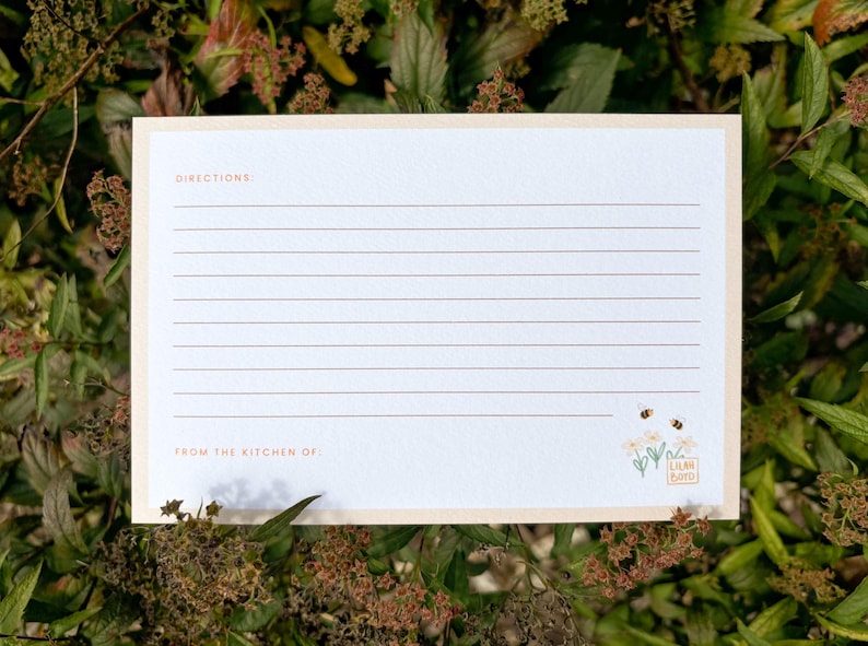 Peut inclure: Une carte de recette avec un fond cr&egrave;me et une bordure p&ecirc;che clair. La carte comporte des lignes horizontales pour l'&eacute;criture, avec les mots "DIRECTIONS" et "FROM THE KITCHEN OF" imprim&eacute;s en marron clair. Une petite illustration florale et d'abeilles est dans le coin inf&eacute;rieur droit.