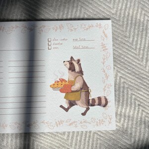 May include: A light blue recipe card with a floral border. The card has lines for writing down ingredients and instructions. There are checkboxes for slow cooker, stovetop, and oven. The card also has spaces for prep time and total time. A cartoon raccoon wearing an apron is holding a pie.