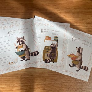 May include: Three printable recipe cards featuring illustrations of raccoons in various kitchen settings. Each card has a checklist for ingredients, a space for prep time, cook time, and total time, and a line for notes. The cards are decorated with a floral border.