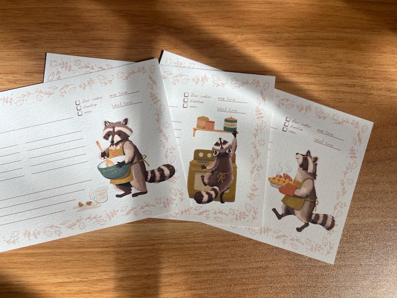 May include: Three printable recipe cards featuring illustrations of raccoons in various kitchen settings. Each card has a checklist for ingredients, a space for prep time, cook time, and total time, and a line for notes. The cards are decorated with a floral border.