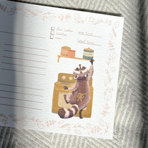 May include: A printable recipe card with a raccoon wearing an apron and holding a jar of sugar. The raccoon is standing in front of a yellow oven. The card has a checklist for slow cooker, stovetop, and oven. It also has spaces to write in prep time and total time.