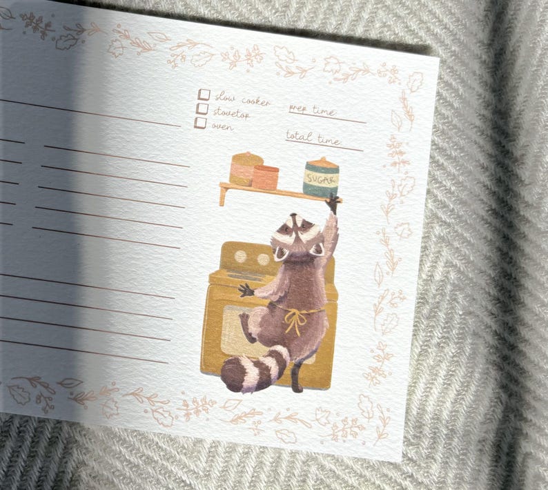 May include: A printable recipe card with a raccoon wearing an apron and holding a jar of sugar. The raccoon is standing in front of a yellow oven. The card has a checklist for slow cooker, stovetop, and oven. It also has spaces to write in prep time and total time.