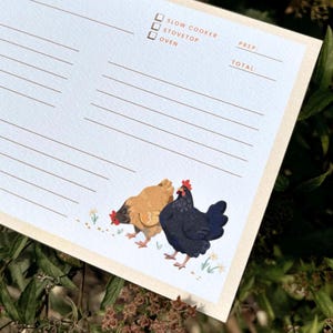 Peut inclure: Un bloc-notes de recettes bleu clair avec une bordure cr&egrave;me. Le bloc-notes pr&eacute;sente un dessin de deux poulets et les mots "Slow Cooker", "Stovetop" et "Oven". Le bloc-notes comporte des lignes pour les ingr&eacute;dients et les instructions de la recette.