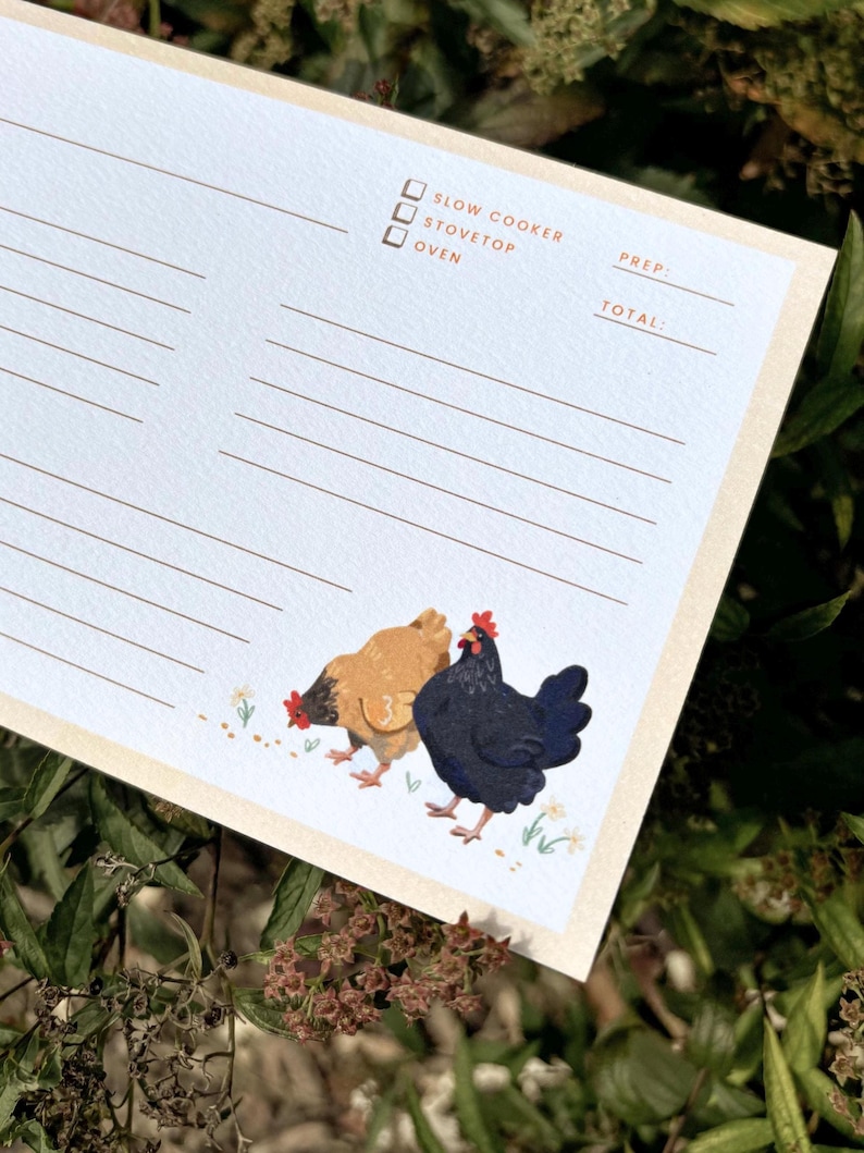 Peut inclure: Un bloc-notes de recettes bleu clair avec une bordure cr&egrave;me. Le bloc-notes pr&eacute;sente un dessin de deux poulets et les mots "Slow Cooker", "Stovetop" et "Oven". Le bloc-notes comporte des lignes pour les ingr&eacute;dients et les instructions de la recette.