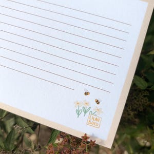Peut inclure: Un bloc-notes lign&eacute; avec une bordure cr&egrave;me. Le bloc-notes pr&eacute;sente une illustration florale avec deux abeilles et le texte "Lilah Boyd" dans un petit carr&eacute;. L'arri&egrave;re-plan est un flou de feuillage vert.