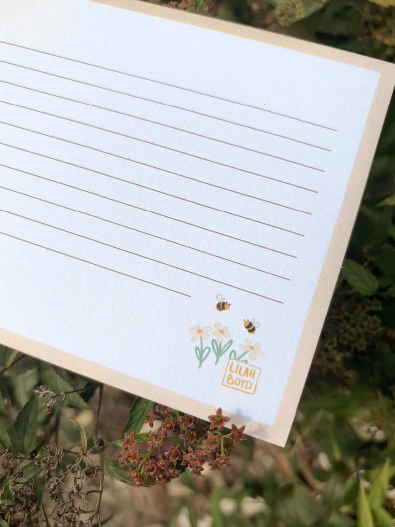 Peut inclure: Un bloc-notes lign&eacute; avec une bordure cr&egrave;me. Le bloc-notes pr&eacute;sente une illustration florale avec deux abeilles et le texte "Lilah Boyd" dans un petit carr&eacute;. L'arri&egrave;re-plan est un flou de feuillage vert.