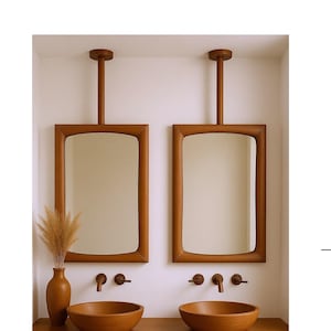 Pode incluir: Uma bancada de casa de banho com dois espelhos com moldura de madeira suspensos do teto. Abaixo estão duas pias de madeira, torneiras e um armário de madeira. Um vaso com plantas secas está sobre a bancada.