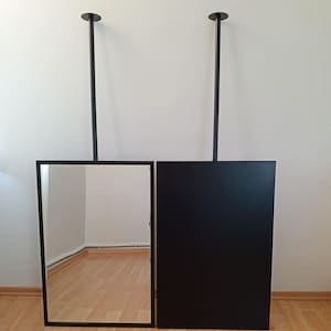 May include: Two black framed mirrors hanging from the ceiling with black metal rods. The mirror on the left reflects the wooden floor and a white wall. The mirror on the right is a solid black surface.