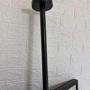May include: A matte black ceiling mount fixture featuring a cylindrical vertical rod attached to a circular base plate. The rod connects to a rectangular frame, providing a sturdy support structure for a mirror or display panel in a vertical orientation.