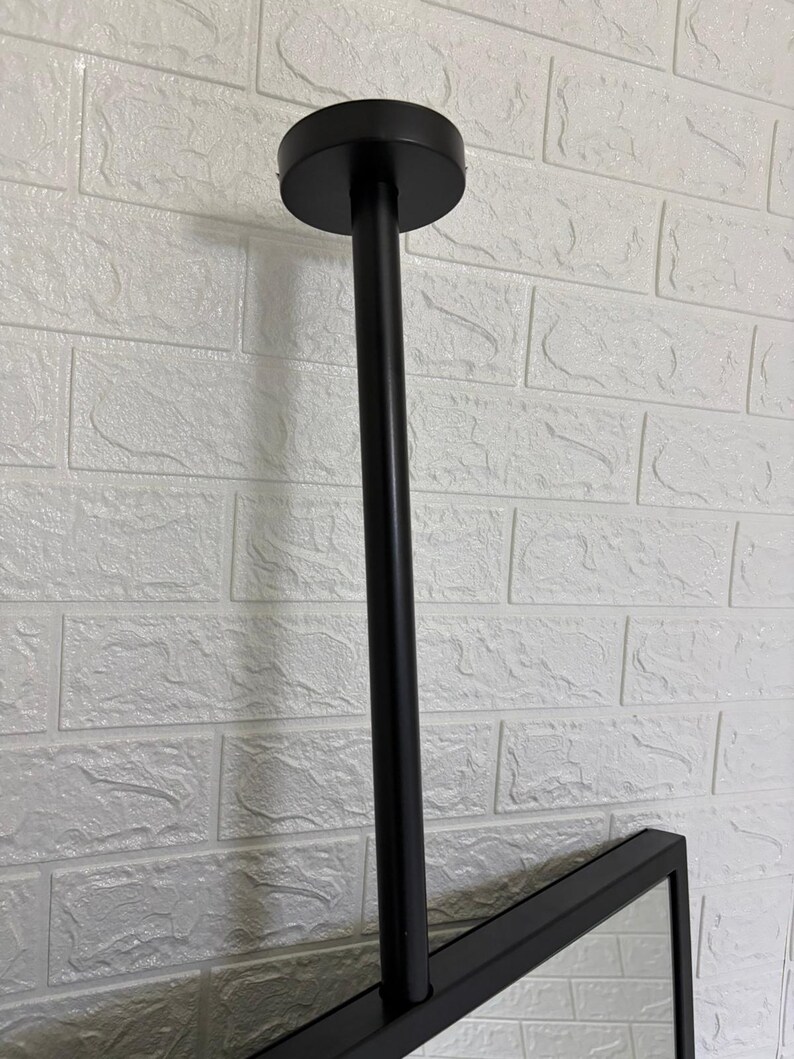 May include: A matte black ceiling mount fixture featuring a cylindrical vertical rod attached to a circular base plate. The rod connects to a rectangular frame, providing a sturdy support structure for a mirror or display panel in a vertical orientation.