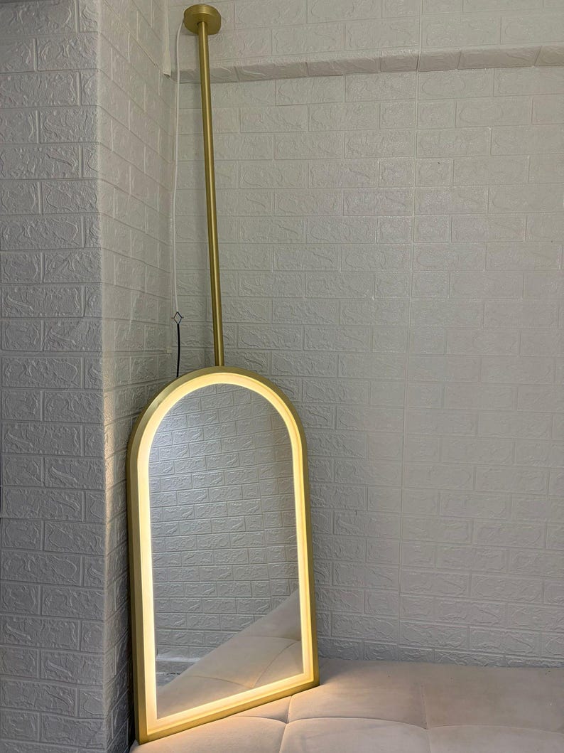 May include: An arched floor mirror with a gold-toned frame and an integrated LED light strip along the inner edge. The mirror is suspended from a vertical gold-toned pole that extends upward to a circular ceiling mount, featuring a thin white power cord.