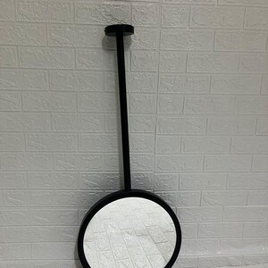 May include: A circular wall mirror featuring a matte black frame and a long, slender black vertical rod extending from the top of the frame to a small circular mounting plate, creating a clean and functional design for wall mounting.