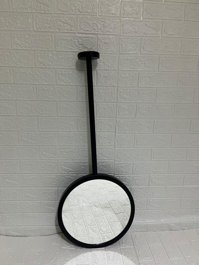 May include: A circular wall mirror featuring a matte black frame and a long, slender black vertical rod extending from the top of the frame to a small circular mounting plate, creating a clean and functional design for wall mounting.