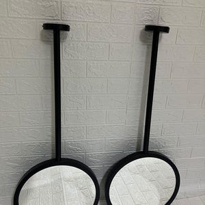 May include: Two circular mirrors with matte black frames, each attached to a long, vertical black metal rod that extends upward to a circular mounting plate. The mirrors feature a clean, reflective glass surface and a simple, uniform design.