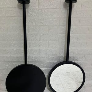 May include: Two circular wall-mounted mirrors with matte black frames and long, slender black stems extending to a circular mounting plate. One mirror shows a reflective glass surface, while the other displays a solid black circular back panel.