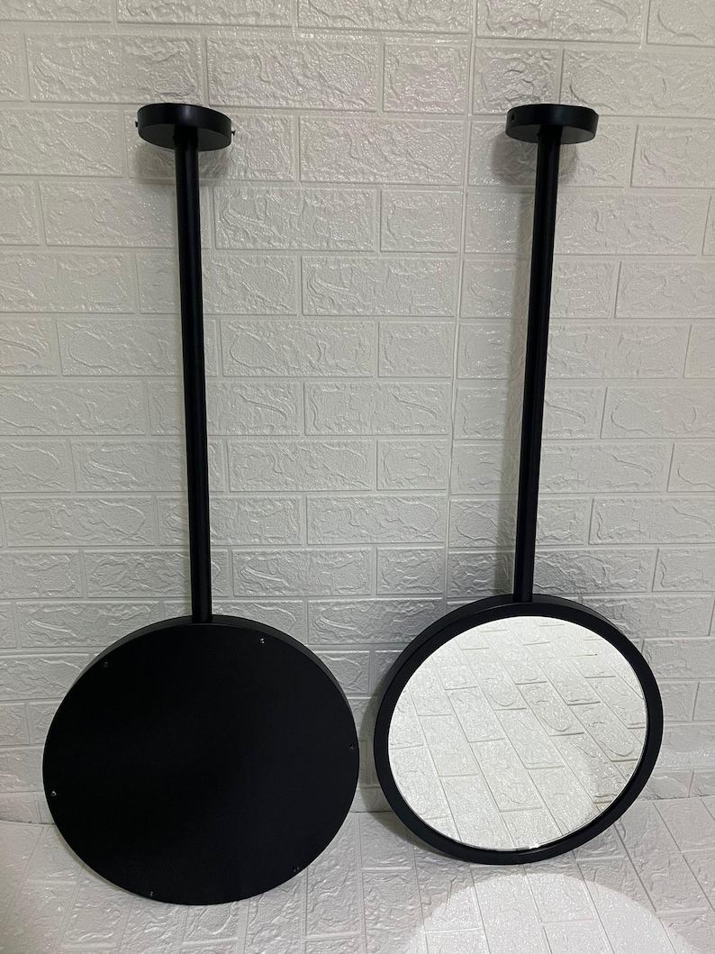 May include: Two circular wall-mounted mirrors with matte black frames and long, slender black stems extending to a circular mounting plate. One mirror shows a reflective glass surface, while the other displays a solid black circular back panel.