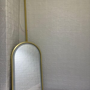 May include: An arched wall mirror with a gold-toned metal frame and an integrated LED light strip along the inner edge. The mirror is suspended from a vertical gold-toned metal rod that attaches to the ceiling with a circular gold mount.