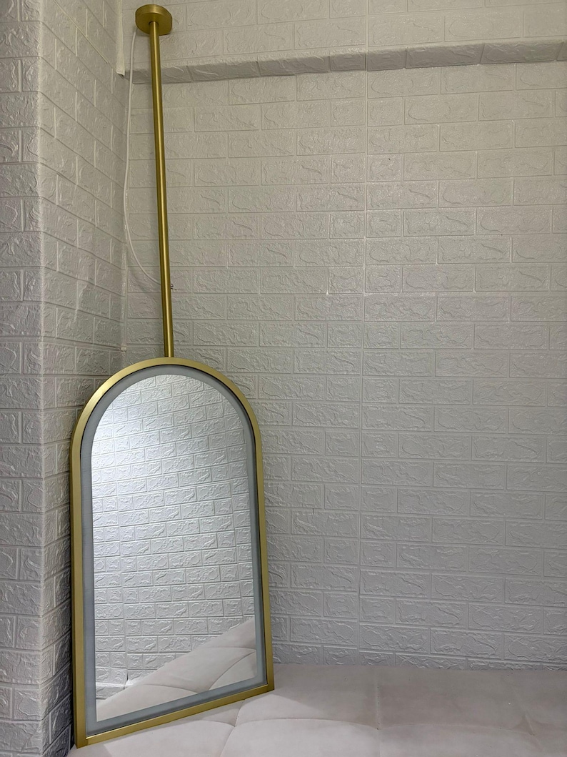 May include: An arched wall mirror with a gold-toned metal frame and an integrated LED light strip along the inner edge. The mirror is suspended from a vertical gold-toned metal rod that attaches to the ceiling with a circular gold mount.