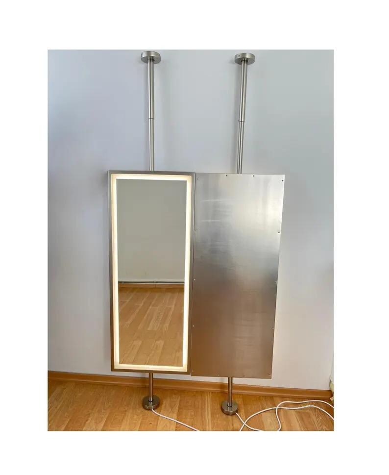 May include: A floor-to-ceiling mounted unit featuring a rectangular mirror with an integrated LED light strip on the left side and a solid brushed metal panel on the right side. The assembly is supported by two vertical silver metal poles.