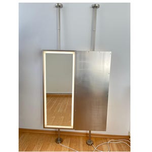 May include: A modern, stainless steel standing mirror with a rectangular, illuminated frame. The mirror is attached to a matching stainless steel panel. The unit is supported by two vertical poles and stands on a wooden floor.