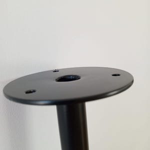 Może przedstawiać: Czarna metalowa noga stołu z okrągłą podstawą i trzema otworami na śruby. Podstawa ma średnicę około 10 cm.