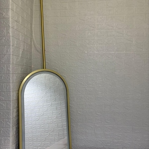 May include: An arched wall mirror with a gold-toned metal frame and an integrated LED light strip along the inner edge. The mirror is suspended from a vertical gold-toned metal rod that attaches to the ceiling with a circular gold mount.