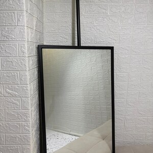 May include: A rectangular mirror with a thin black metal frame, suspended from a single vertical black metal rod that attaches to a circular ceiling mount. The mirror surface is clear and reflective, showing a portion of a light-colored surface.