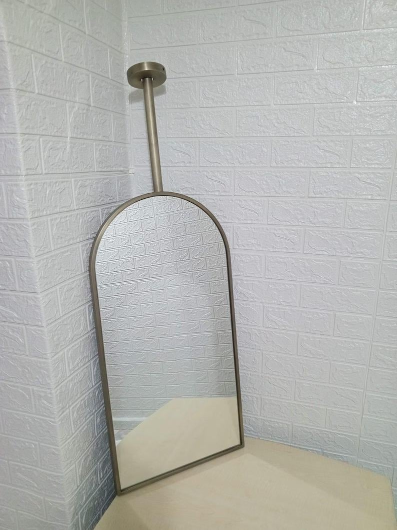 May include: An arched wall mirror with a metallic bronze frame. The mirror features a vertical rod extending from the top center of the arch to a circular mounting plate, designed for ceiling or wall installation, creating a sleek, elongated look.