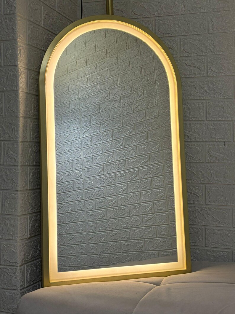 May include: An arched floor mirror featuring a metallic gold frame and an integrated LED light strip that follows the curved top and straight sides, providing a bright, even glow around the reflective glass surface of the tall mirror.