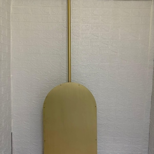 May include: A gold-colored metal wall mirror with an arched top shape. The mirror features a long, vertical gold metal rod extending from the top center of the arch to a circular mounting plate at the top, creating a suspended appearance.