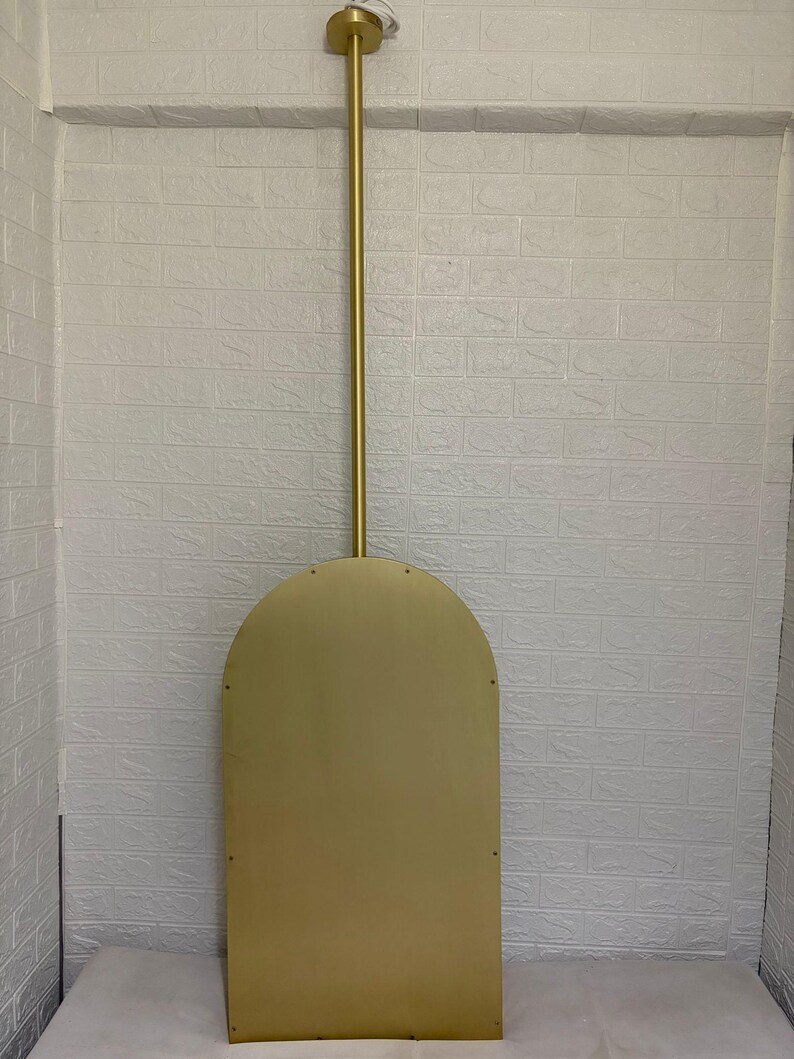 May include: A gold-colored metal wall mirror with an arched top shape. The mirror features a long, vertical gold metal rod extending from the top center of the arch to a circular mounting plate at the top, creating a suspended appearance.