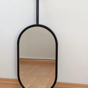 May include: An oval-shaped mirror with a smooth black frame. The mirror is attached to a vertical black metal rod that extends upward, ending in a circular mounting plate designed for ceiling or wall installation, creating a suspended appearance.