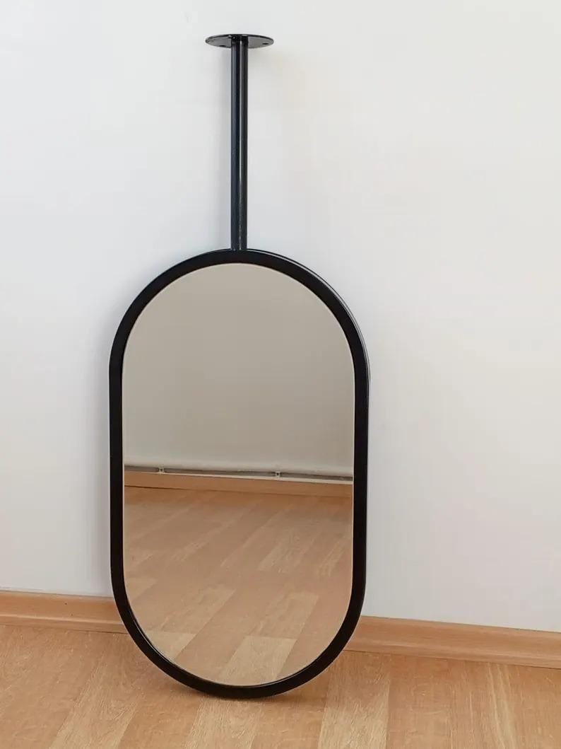 May include: An oval-shaped mirror with a smooth black frame. The mirror is attached to a vertical black metal rod that extends upward, ending in a circular mounting plate designed for ceiling or wall installation, creating a suspended appearance.