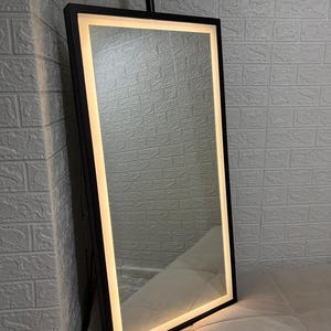 May include: A rectangular floor mirror featuring a slim black frame and an integrated LED light strip that runs along the inner perimeter. The mirror emits a warm, soft glow, illuminating the reflective surface within the dark metal border.