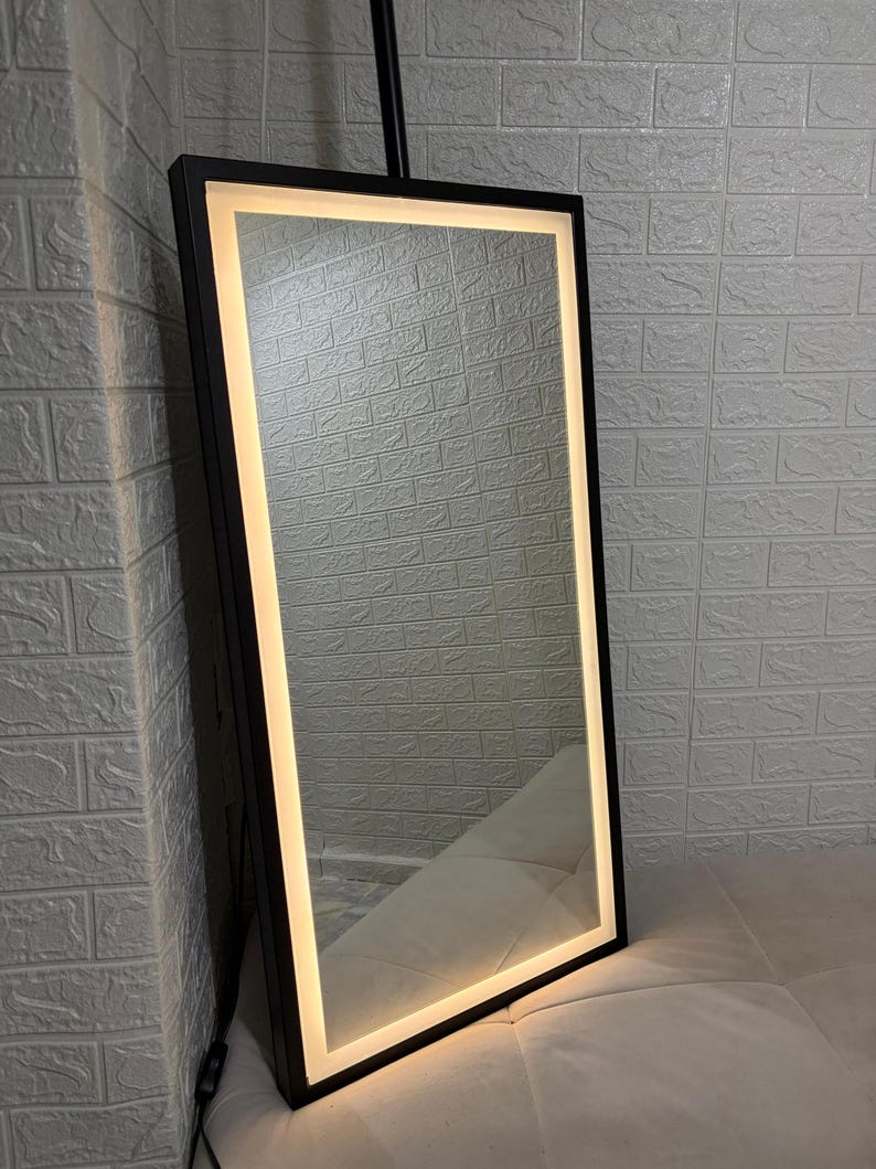May include: A rectangular floor mirror featuring a slim black frame and an integrated LED light strip that runs along the inner perimeter. The mirror emits a warm, soft glow, illuminating the reflective surface within the dark metal border.
