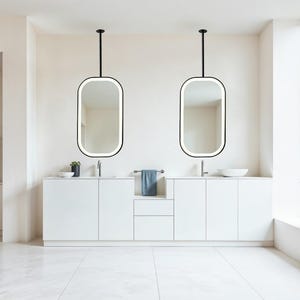 May include: A modern bathroom with two oval mirrors suspended from the ceiling. The mirrors have black frames and integrated lighting. White cabinets and a blue towel add contrast to the neutral color scheme. A small plant and a stool are also visible.