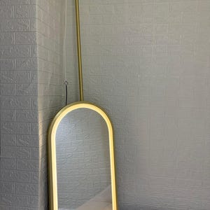 May include: An arched floor mirror with a gold-toned metal frame and an integrated LED light strip along the inner edge. The mirror is suspended from a vertical gold-toned metal rod that extends upward to a circular ceiling mount fixture.