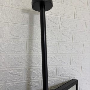 May include: A matte black ceiling mount fixture featuring a cylindrical vertical rod attached to a circular base plate. The rod connects to a rectangular frame, providing a sturdy support structure for a mirror or display panel in a vertical orientation.