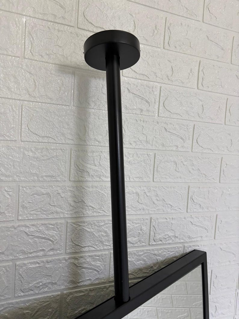 May include: A matte black ceiling mount fixture featuring a cylindrical vertical rod attached to a circular base plate. The rod connects to a rectangular frame, providing a sturdy support structure for a mirror or display panel in a vertical orientation.