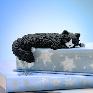 Puede incluir: Un marcapáginas con forma de gato blanco y negro descansa sobre una pila de libros azul claro. El gato está acurrucado, pareciendo dormir. Los libros tienen diseños de copos de nieve.