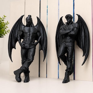 Op de afbeelding: Twee zwarte matte beeldjes die gevleugelde mensachtige figuren met een gespierd postuur voorstellen, gekleed in broeken en laarzen. Elk figuur heeft grote, gedetailleerde vleermuisachtige vleugels die vanaf de rug uitsteken.