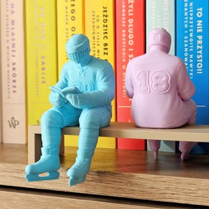 Op de afbeelding: Twee decoratieve beeldjes van hockeyspelers, één lichtblauw en één roze, zitten op een houten plank. De blauwe figuur leest een boek, terwijl de roze figuur nummer 18 op de rug heeft. Ze staan voor een rij kleurrijke boeken.