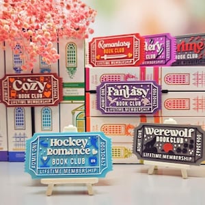May include: A collection of rectangular enamel pins shaped like admission tickets, each featuring unique book club themes such as Romantasy, Fantasy, Hockey Romance, and Werewolf, displayed on small white easels against a backdrop of book spines.