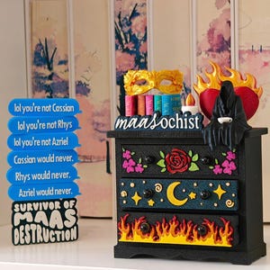 Pode incluir: Uma cómoda preta de três gavetas com motivos florais, celestiais e de chamas. No topo, há uma máscara dourada, livros em miniatura, uma vela e uma figura preta encapuzada com um coração vermelho e chamas. Uma placa azul contém o texto "lol you're not Cassian" e "SURVIVOR OF MAAS DESTRUCTION".